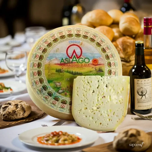 Wheel of Asiago Fresco D.O.P. cheese with a wedge cut, bread rolls, red wine bottle, and pasta dish on a white tablecloth